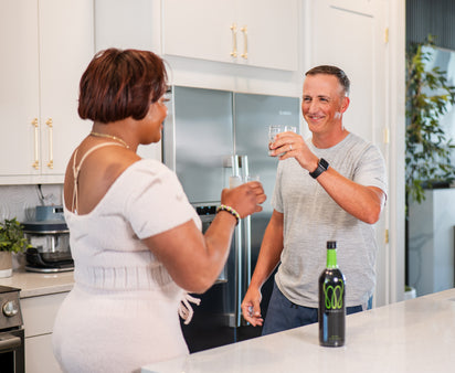 A man and woman smiling and clinking glasses of Monavie Active in a modern kitchen.