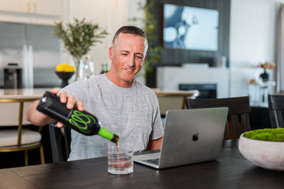 A man pouring a glass of Monavie Active at a dining table while working on a laptop in a modern home office setting.